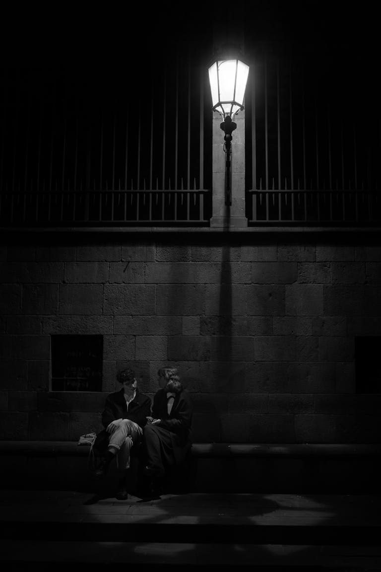 A couple sitting under a streetlight in a black and white urban night scene.