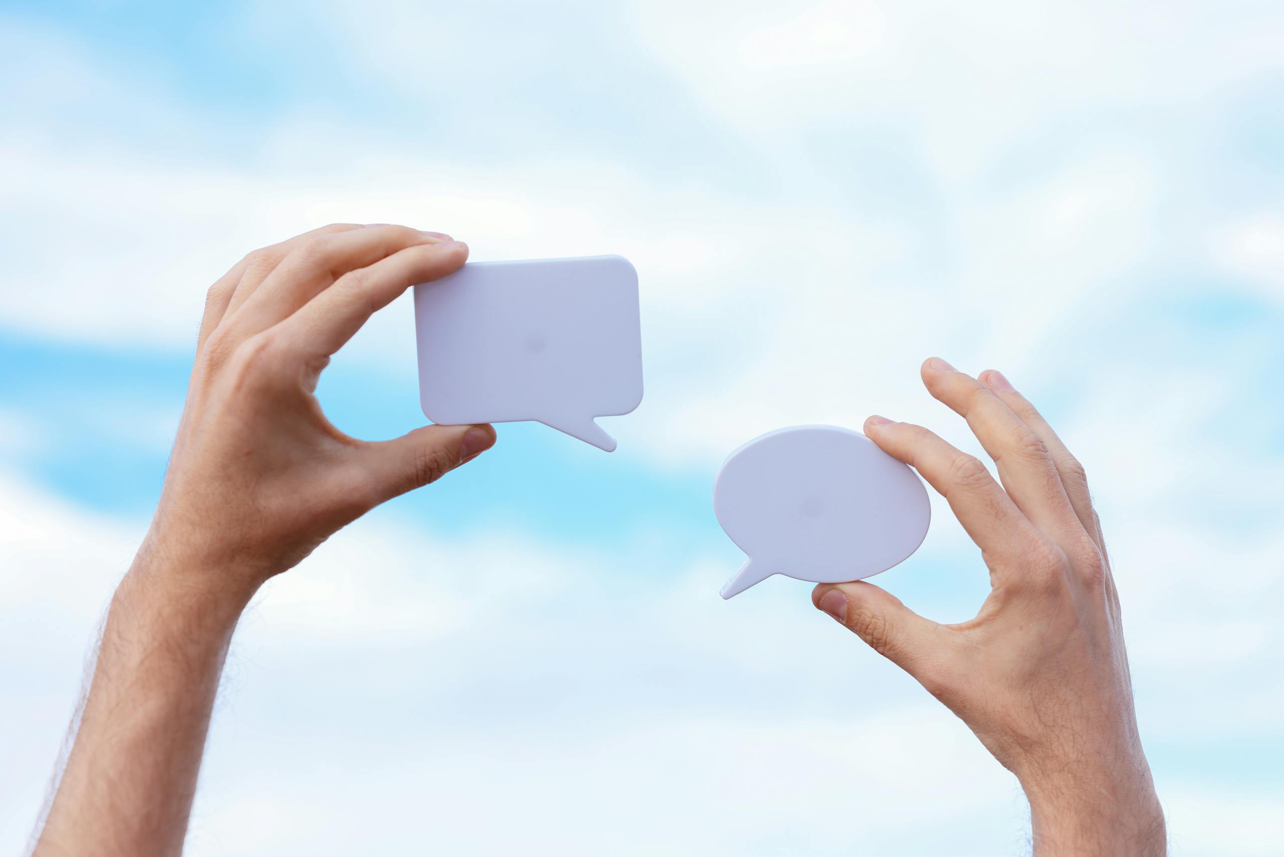 Close-up of hands holding speech bubbles, symbolizing communication and dialogue against a blue sky.