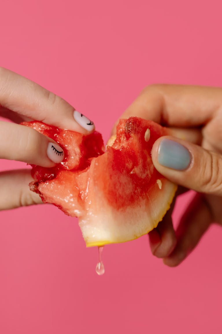 Close-up of hands with manicured nails holding a slice of juicy watermelon against a pink background.