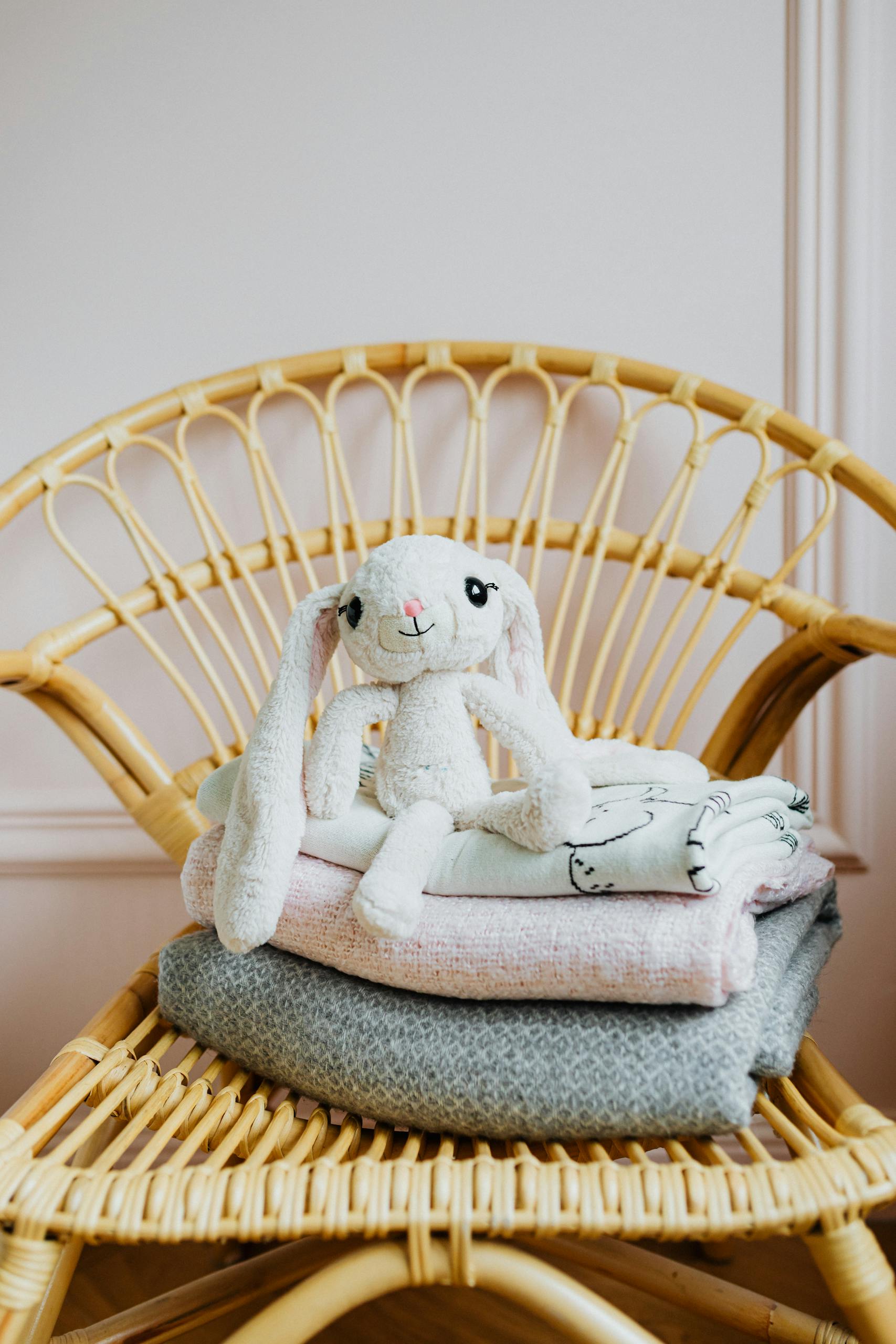 Cozy plush bunny toy sitting on folded blankets on a rattan chair.