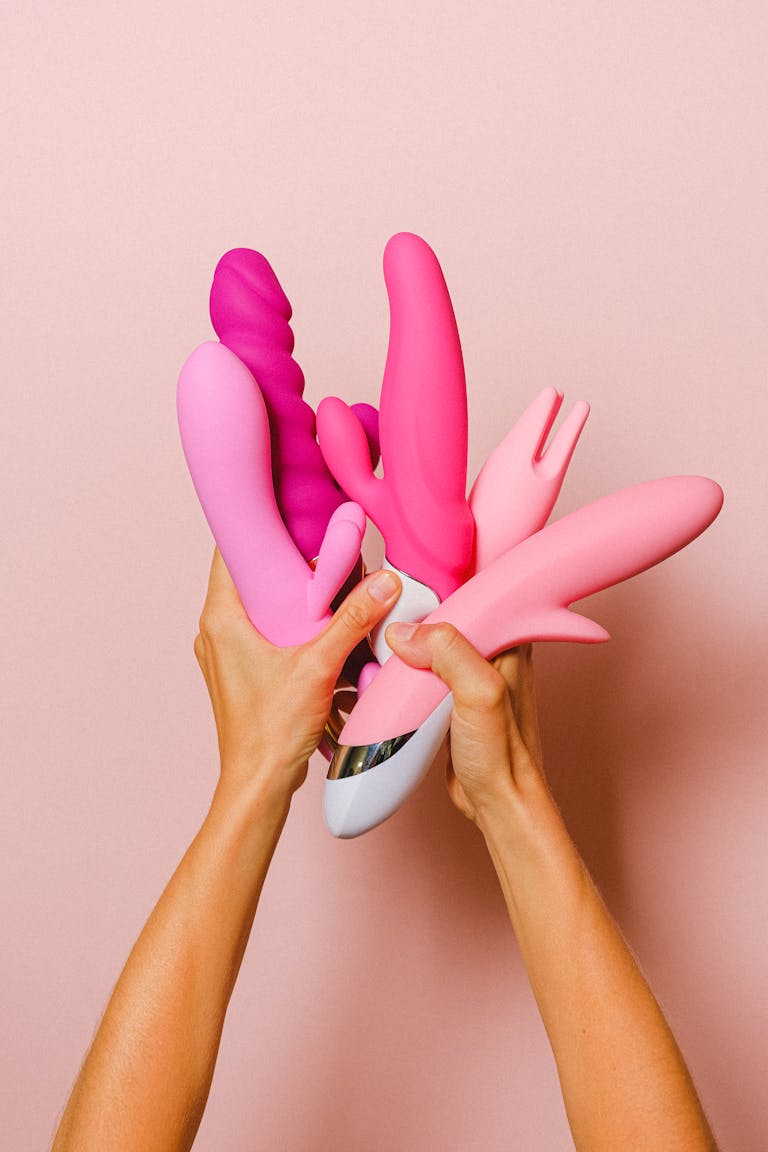 Hands holding various pink silicone vibrators against a pastel pink backdrop.